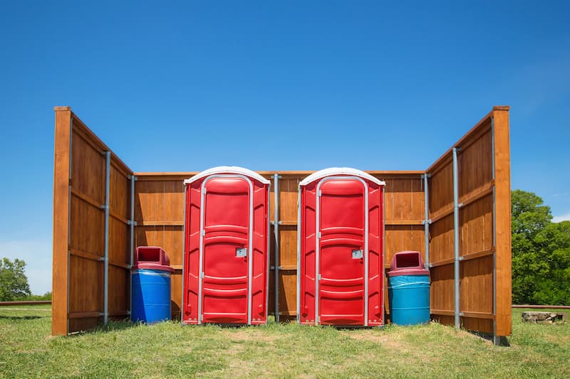 Red portable toilets set up behind a wood privacy fence with waste bins for a festival